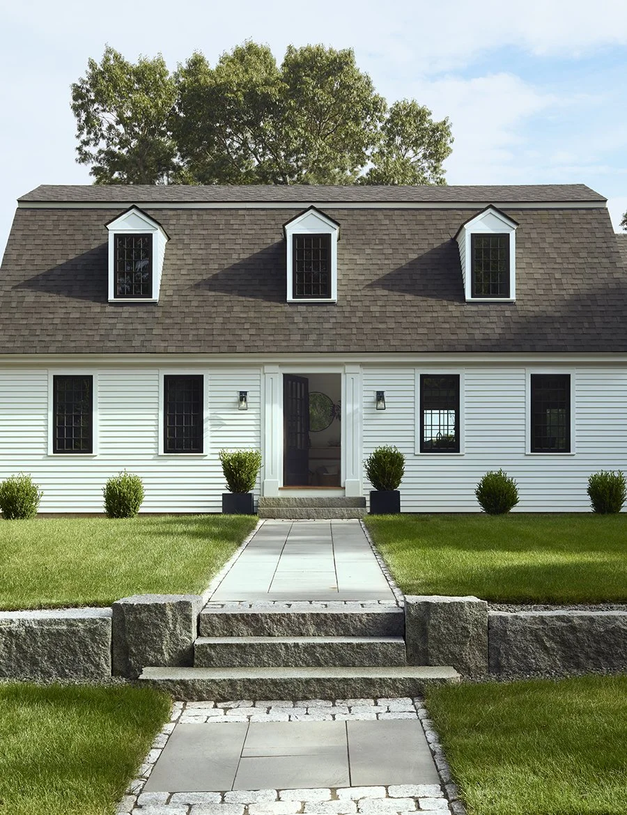 Front of a white house with black windows, a dark shingle roof with three dormer windows, a walkway leading up to the front door, surrounded by green grass and small bushes, with trees in the background.