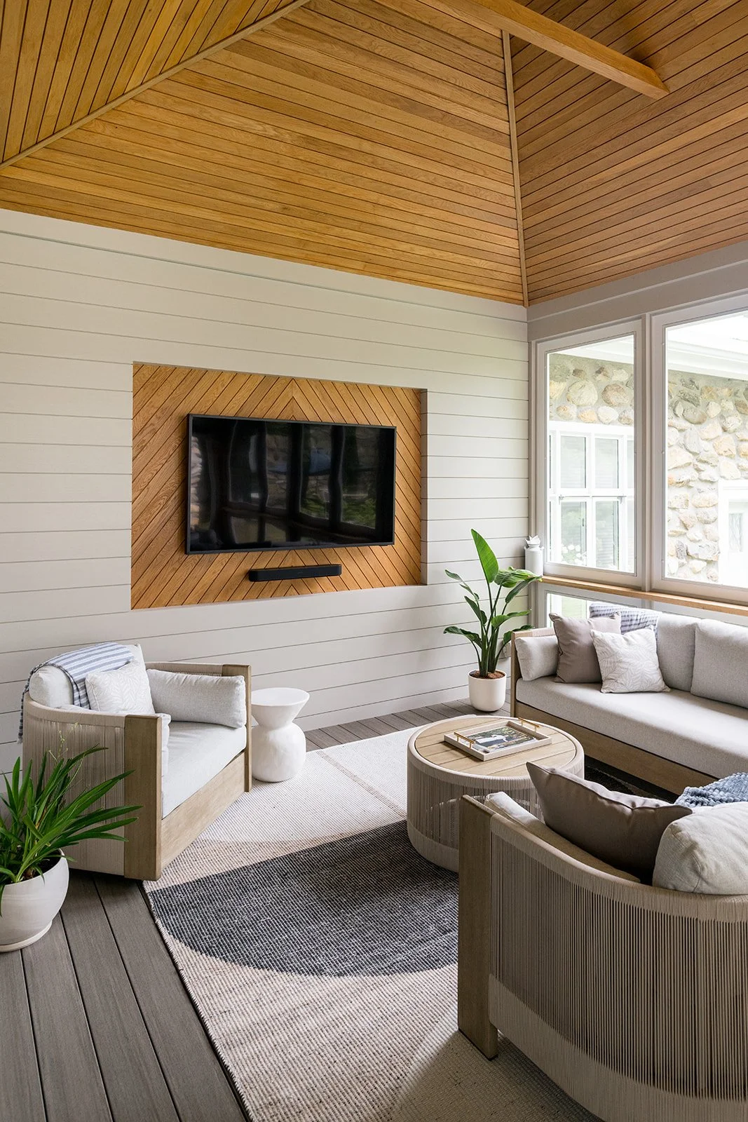 Living room with white and beige furniture, a wooden coffee table, houseplants, a wall-mounted TV with wood accents, large windows, and wood-paneled ceiling and flooring.