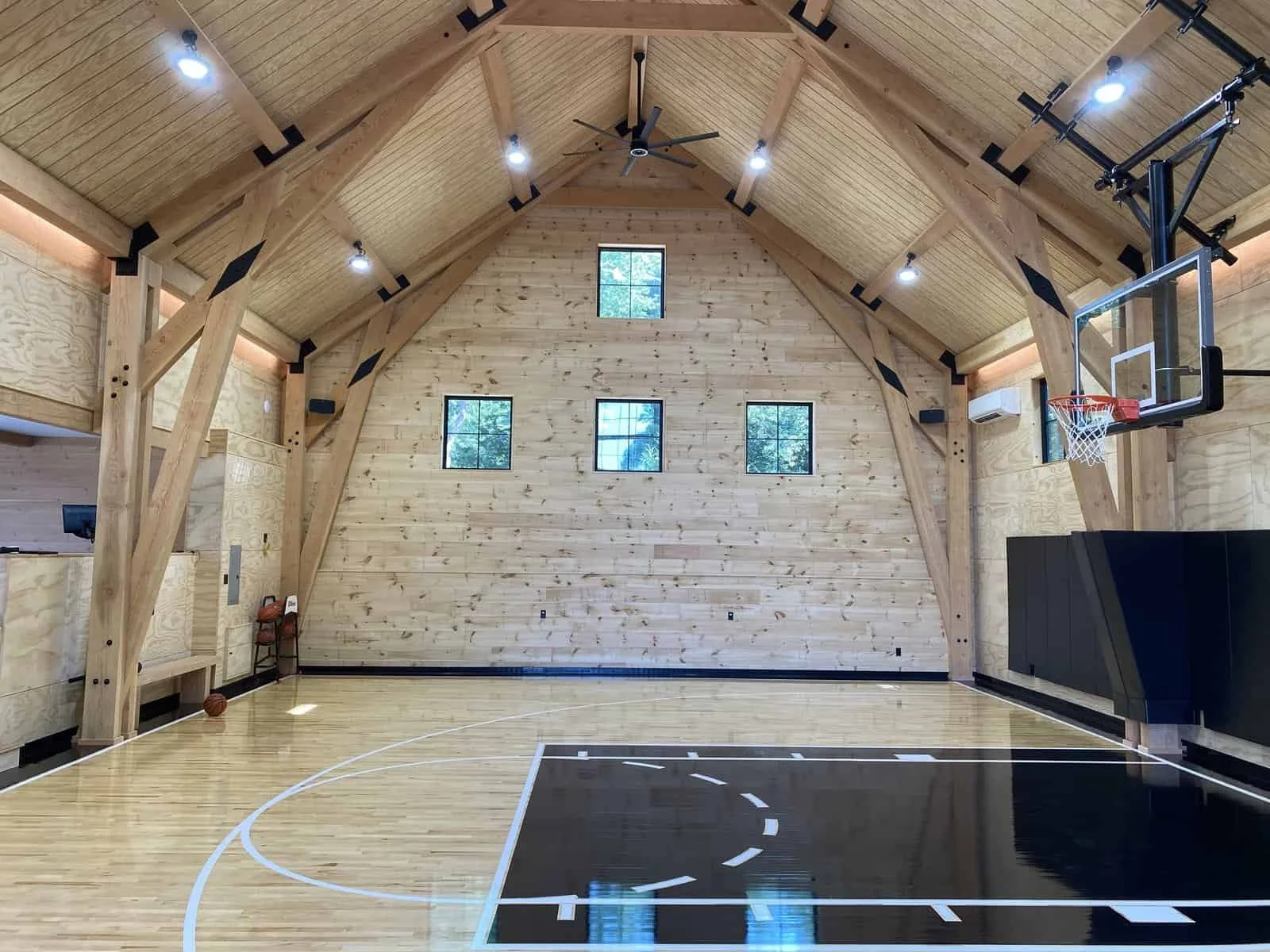 Interior view of a wooden basketball gymnasium with high ceilings, large windows, a mounted basketball hoop, and a black padded wall below the hoop.