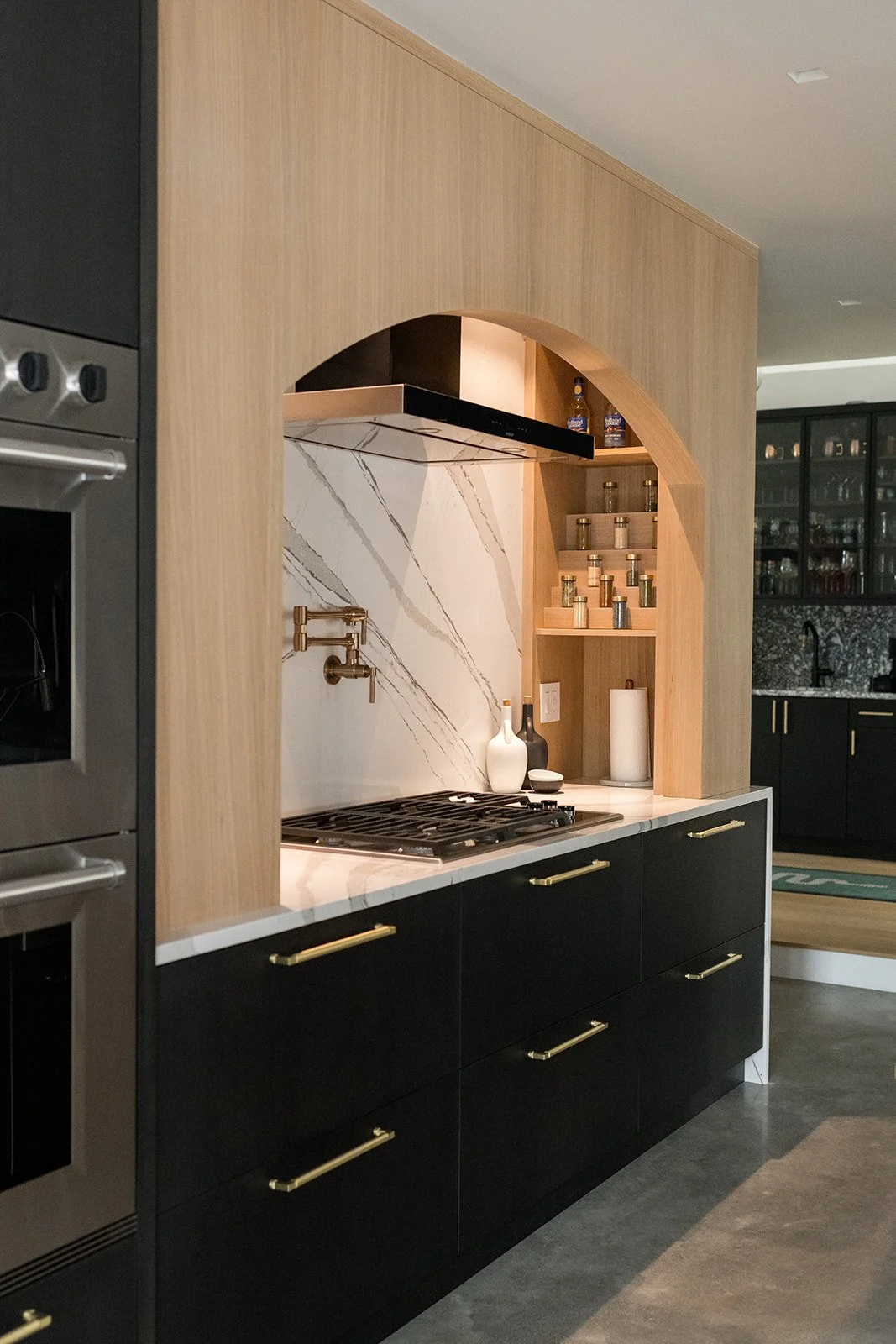Modern kitchen with black cabinets, gold handles, marble countertop, open shelving with spices, and decorative vases.