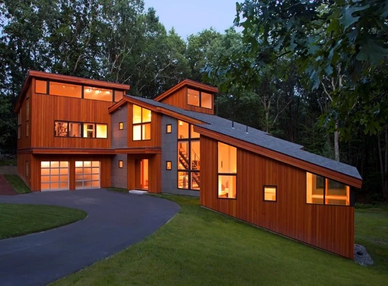 Modern, contemporary multi-story house with a wood facade, large windows, and a sloped roof, illuminated from within at dusk, surrounded by trees and a grassy yard.
