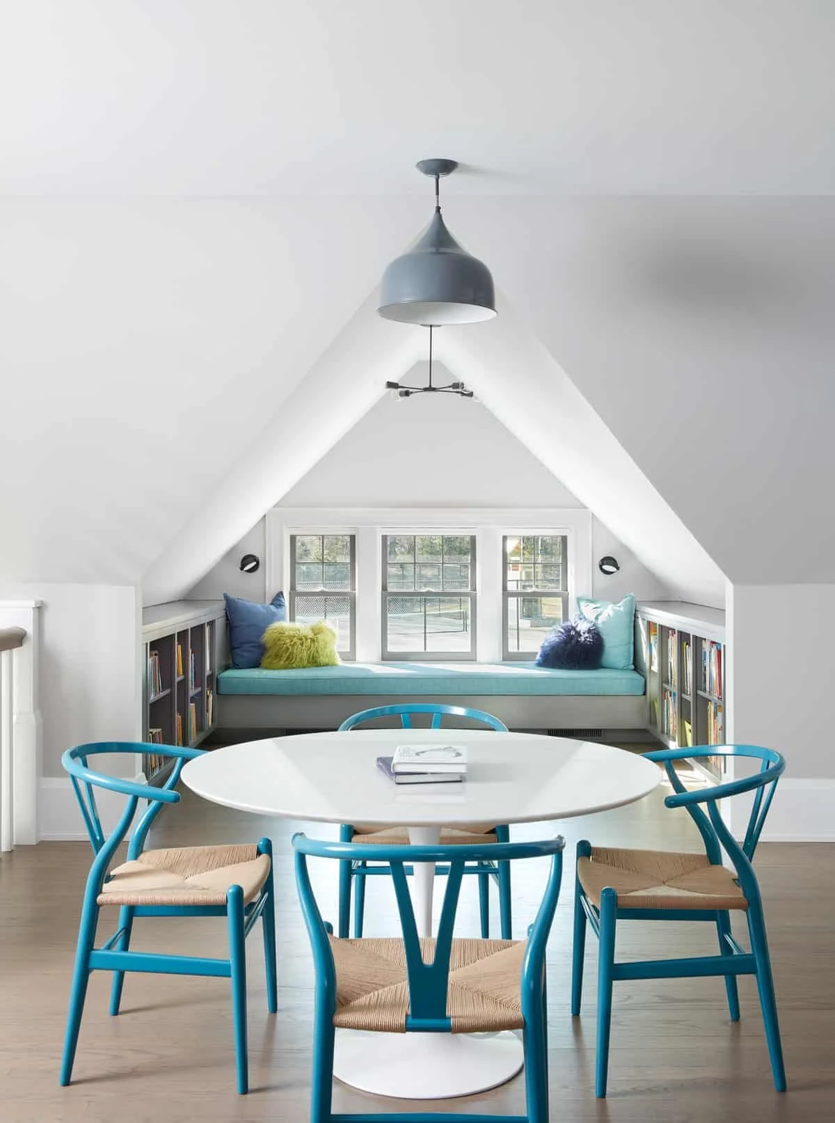 A bright attic room with a sloped ceiling, a window seat with pillows, a round white table with blue chairs, and a black ceiling fan.