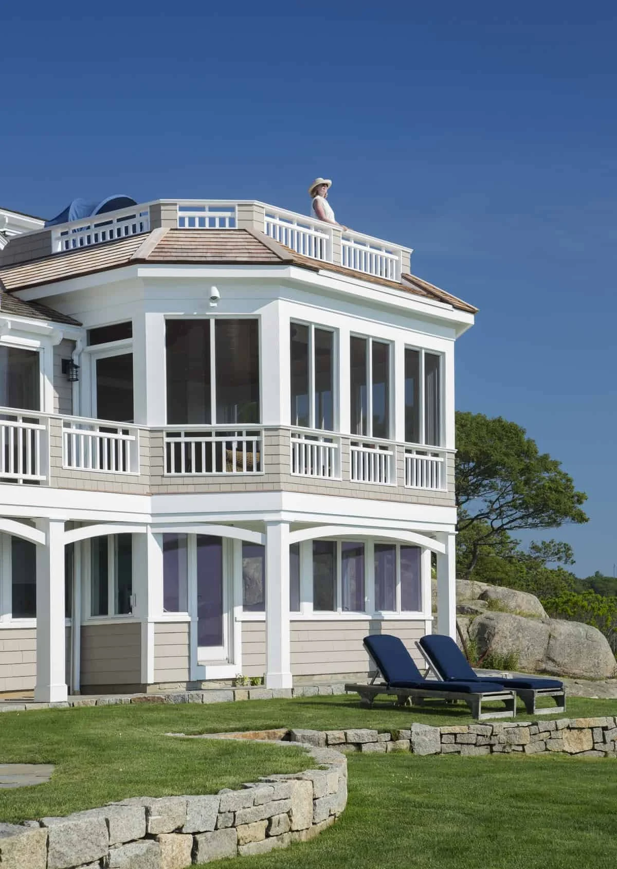 Large white house with multiple levels, balconies, and large windows, situated outdoors on a grassy area with lounge chairs and trees in the background, under a clear blue sky. A woman wearing a hat is on the upper balcony, looking out.