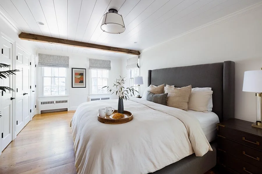 Bright bedroom with a large bed featuring a gray upholstered headboard, topped with beige and gray pillows. There are two nightstands with white lamps on each side. The room has white walls, wooden floors, three windows with gray Roman shades, and a 