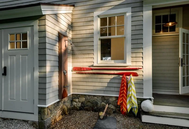 Exterior of a house with gray siding, white window and door trim, a white door with small windows, and a porch with steps. An outdoor shower with a wooden panel and a metal showerhead is beside the window. Red and yellow striped towels hang on hooks,