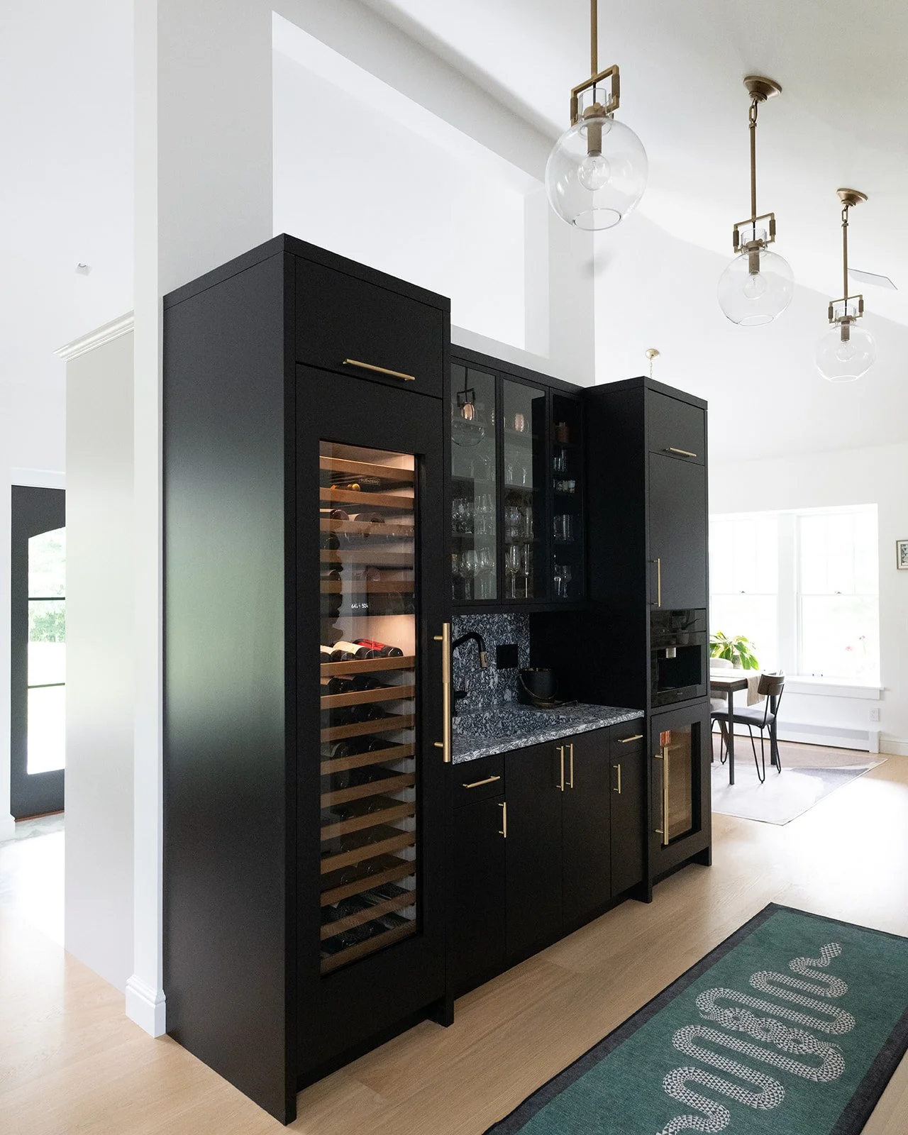Modern black kitchen cabinet with glass doors, wine refrigerator, and black granite countertop in a bright home interior with pendant lights and large windows.