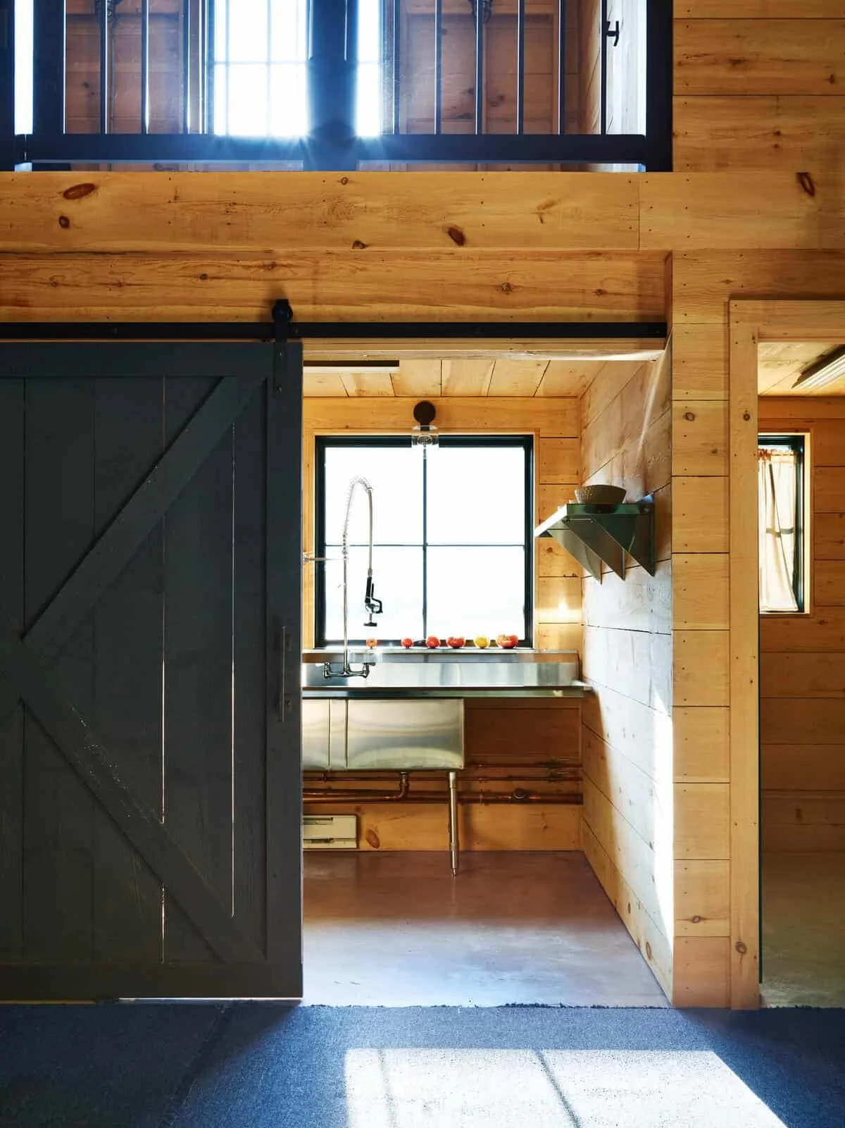 Interior view of a rustic kitchen with wooden walls and floors, a large window above a stainless steel sink, black sliding barn door, and a small shelf with bowls, with sunlight streaming in.