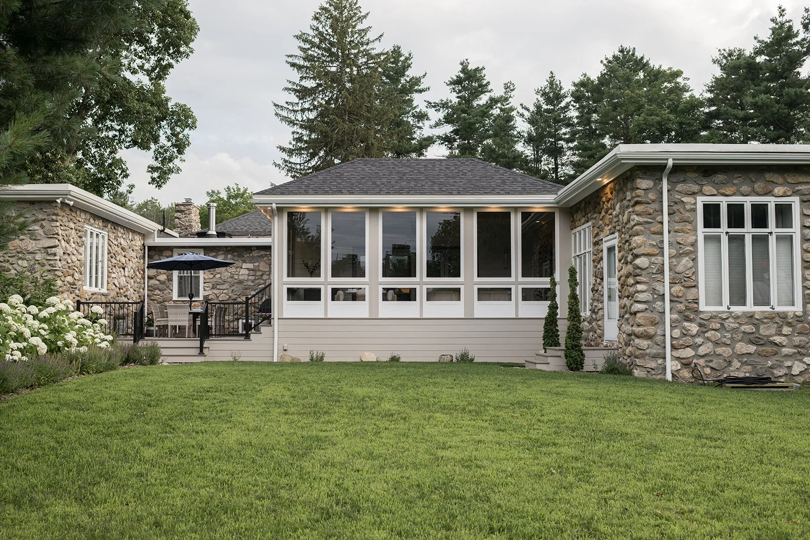 The image shows the backyard of a house with a green lawn, stone walls, and several windows. There is a small patio area with a table, chairs, and an umbrella.