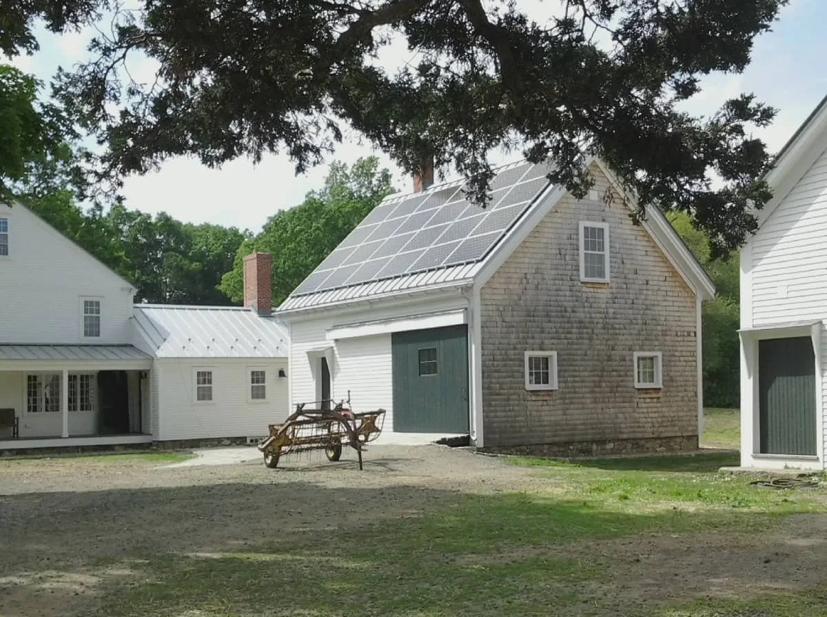 A rural property with a barn that has solar panels on its roof, a white house and a wooden farm tool in the foreground, trees in the background, and a shaded area in the front yard.