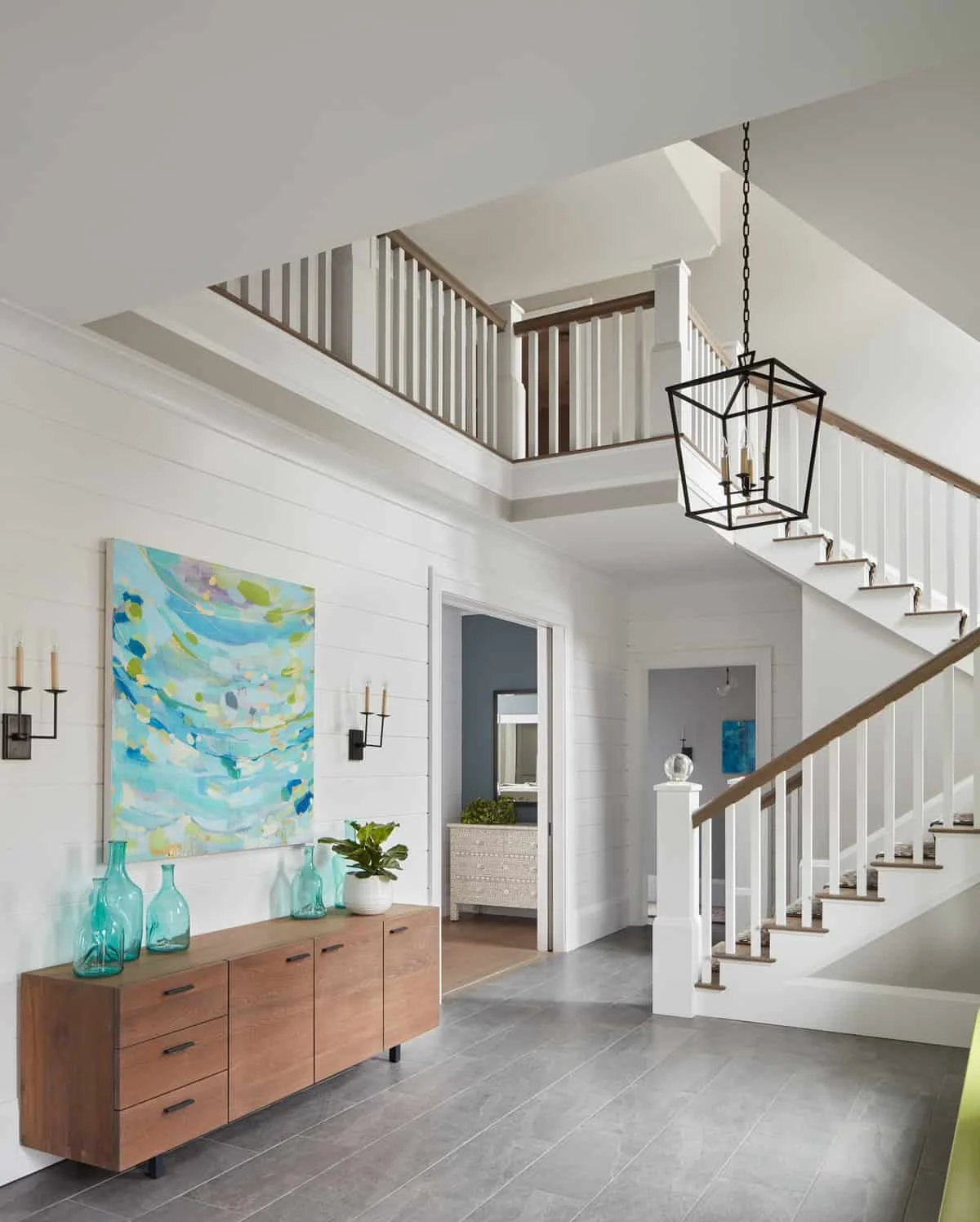 Interior view of a modern house entryway with white walls, wooden flooring, a wooden sideboard with decorative vases, abstract artwork, and a staircase with white railings, a hanging black metal chandelier, and a second-floor balcony.