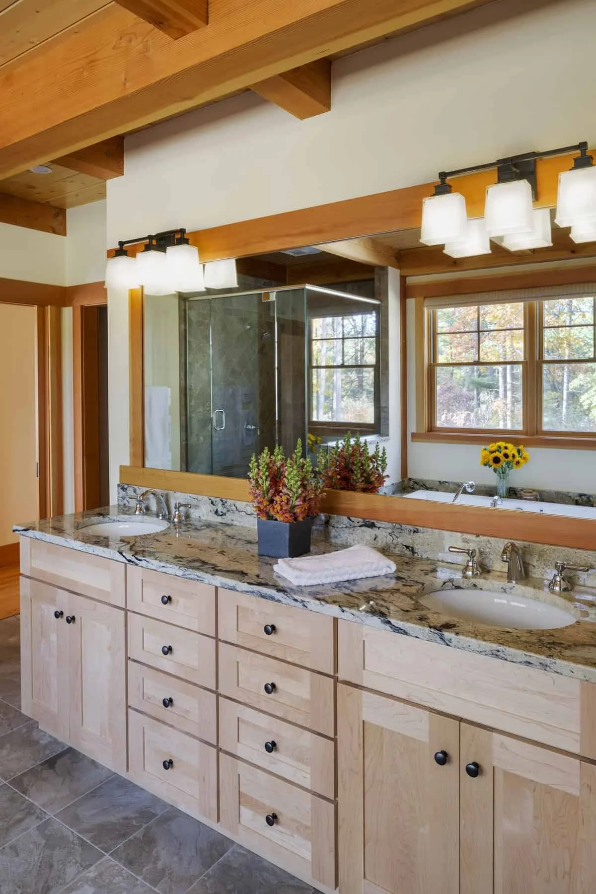 Bathroom vanity with double sinks and a marble countertop, decorated with potted plants and a towel, large mirror, wooden cabinets, and windows showing trees outside.