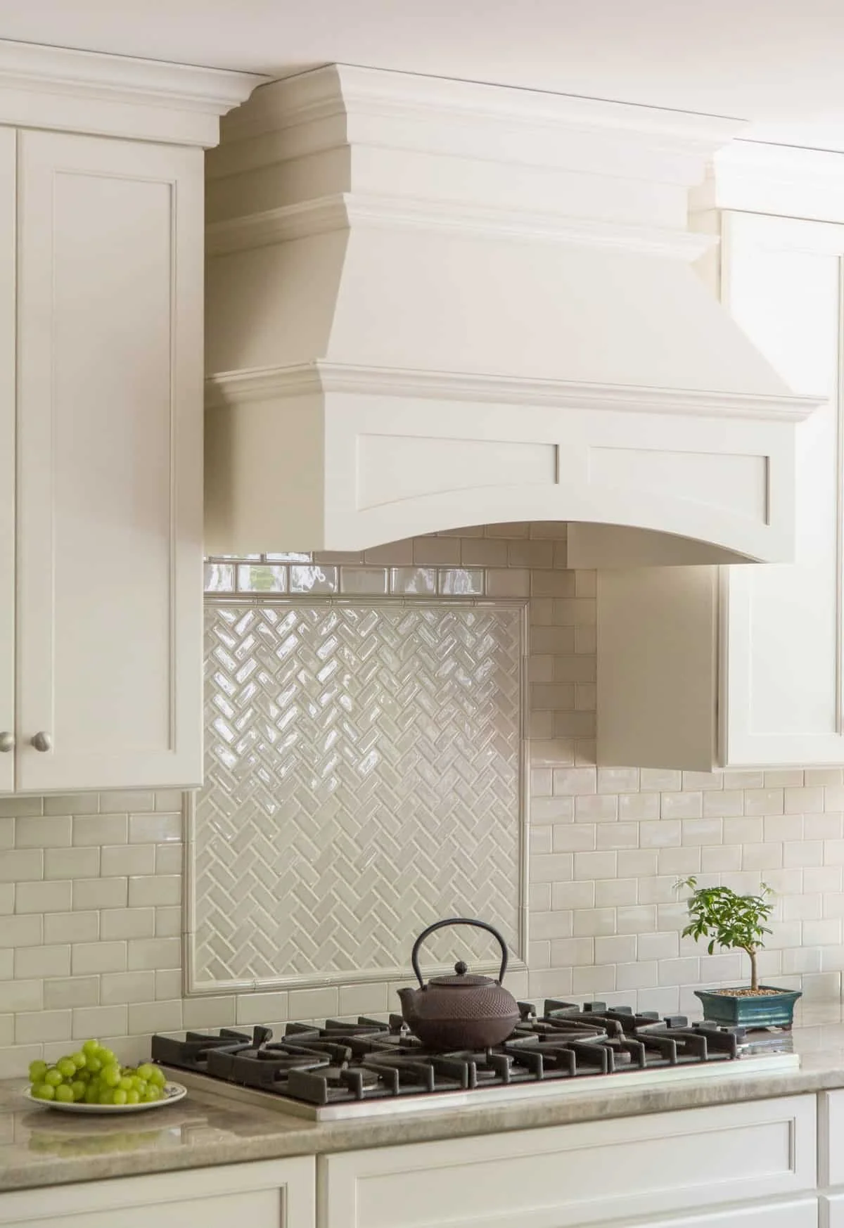 Kitchen with white cabinetry, a gas stove with a black teapot, a small potted plant, grapes on a plate, and a decorative tile backsplash behind the stove.