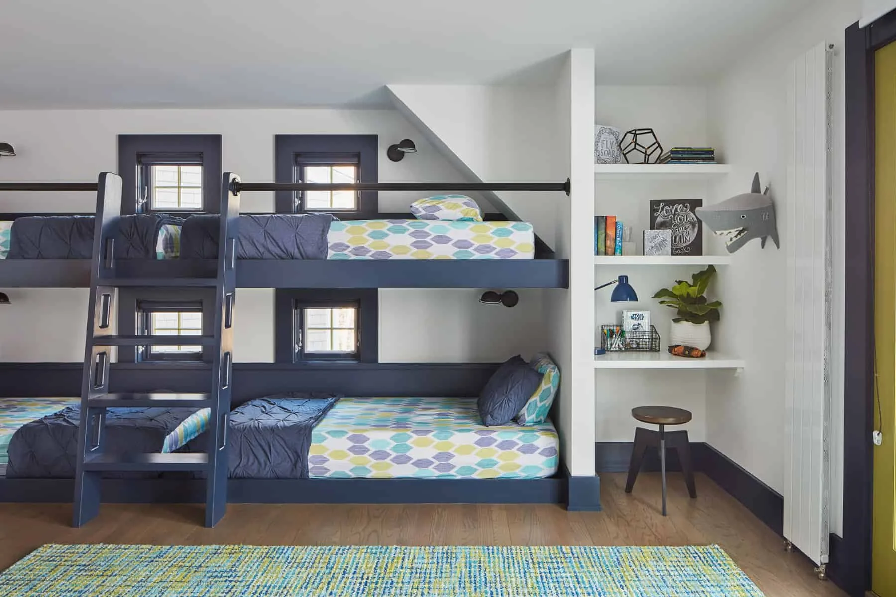 A bedroom with a bunk bed painted in dark gray, featuring two twin mattresses with geometric-patterned sheets and pillows. The top bunk has a ladder for access, and there are small windows behind the bed. To the right, a white wall with built-in shel