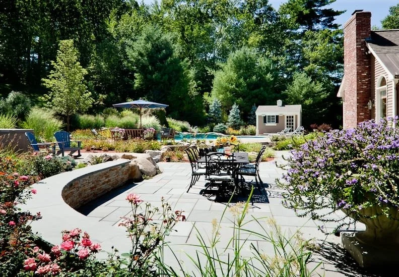 A backyard patio with seating, a small pool, surrounded by lush greenery, flowering plants, and trees, with a small shed and a brick house with a chimney.