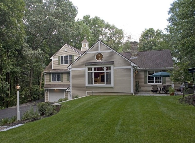 A house with beige siding, multiple gabled roofs, and large windows, situated on a grassy lawn with trees in the background.