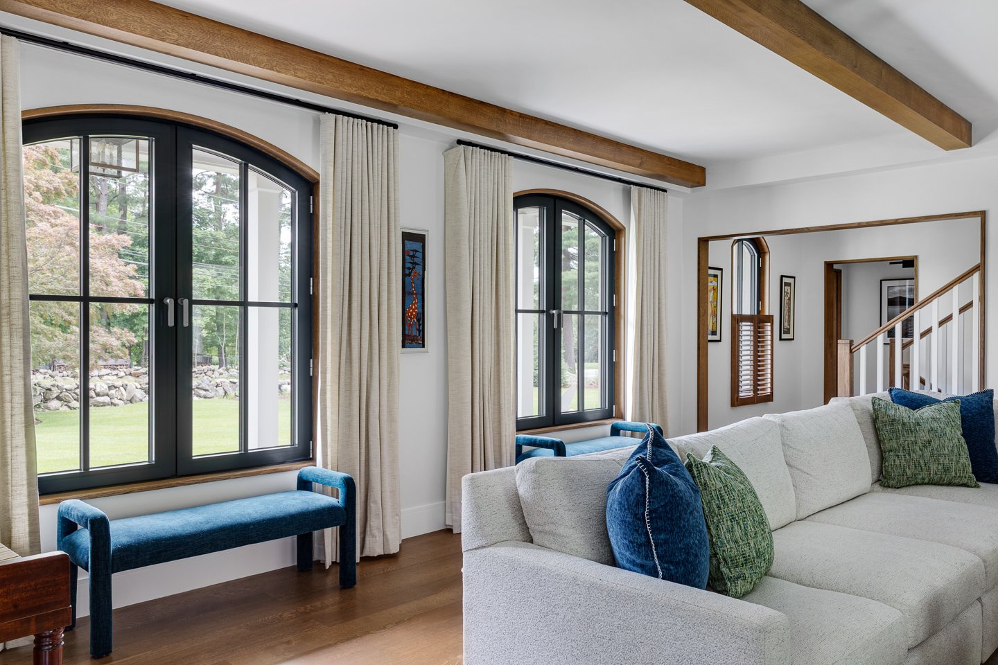 A cozy living room with three large arched windows with black frames and panes, beige curtains, a white sofa with blue and green pillows, wooden ceiling beams, and wooden accents around the windows and staircase. Green trees are visible outside.