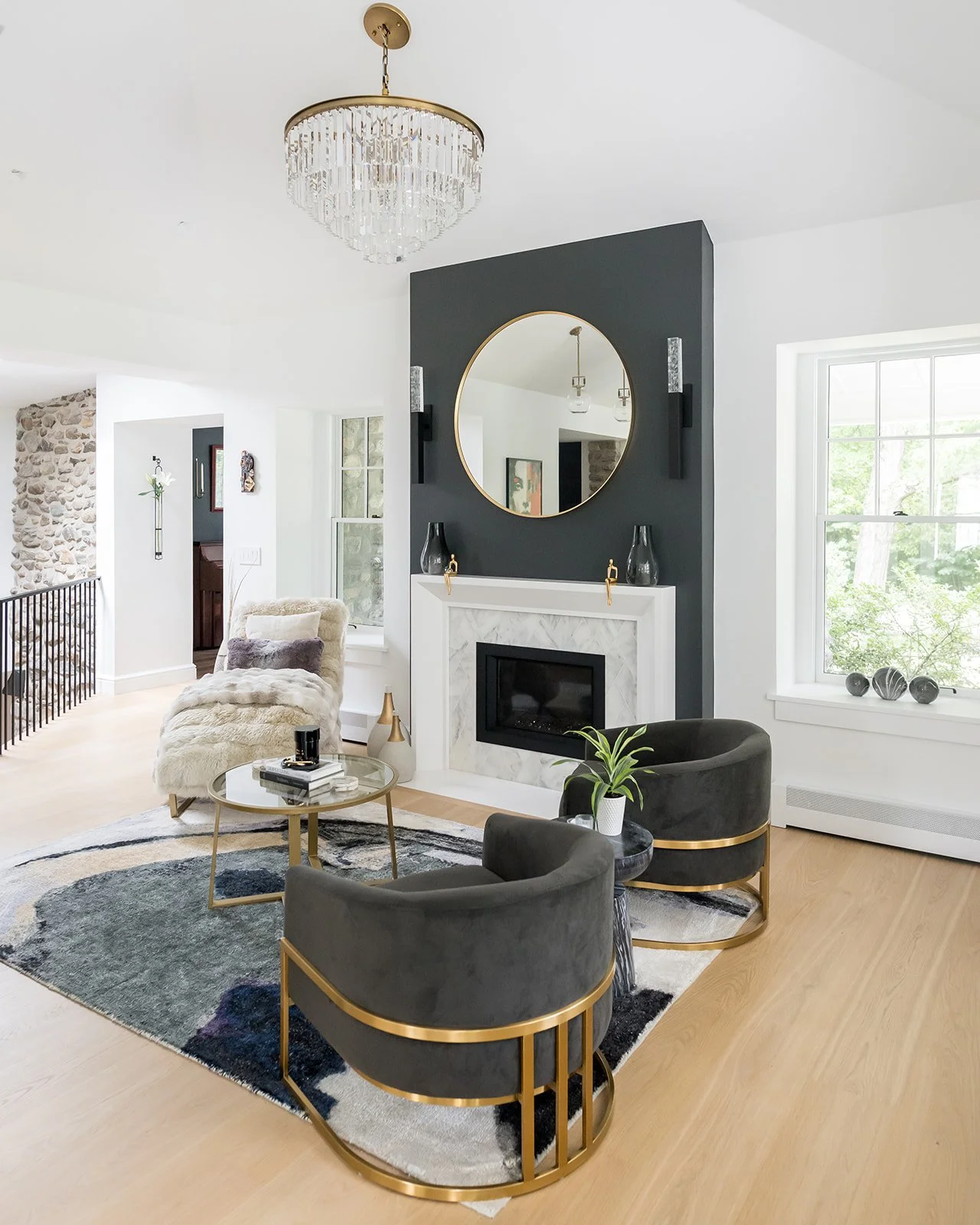 Aesthetic living room with a modern fireplace, round mirror, black and gold armchairs, a fur-covered chair, and a glass-top coffee table, decorated in neutral and black tones with gold accents.
