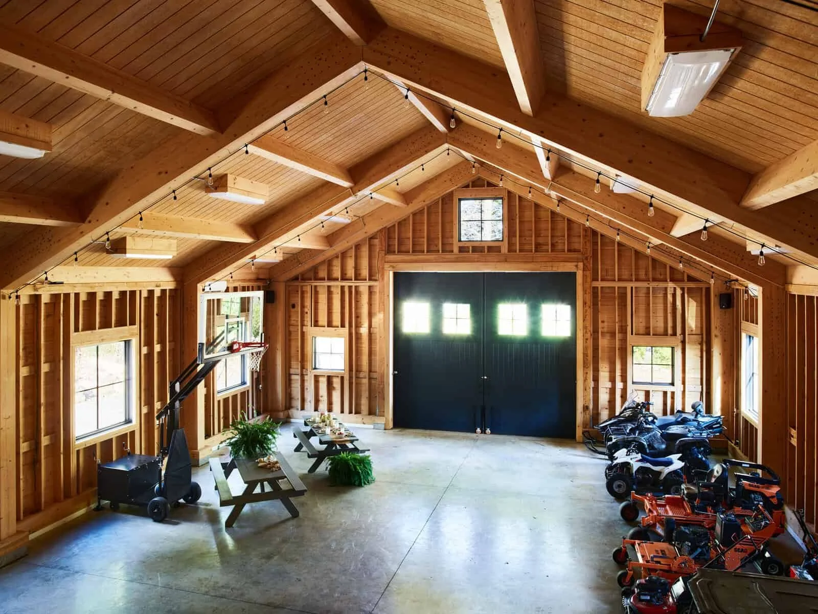 Inside a spacious wooden garage with a high, vaulted ceiling. The garage has multiple windows and contains a basketball hoop, tables with decorations, and several small vehicles or motorcycles parked along the wall.