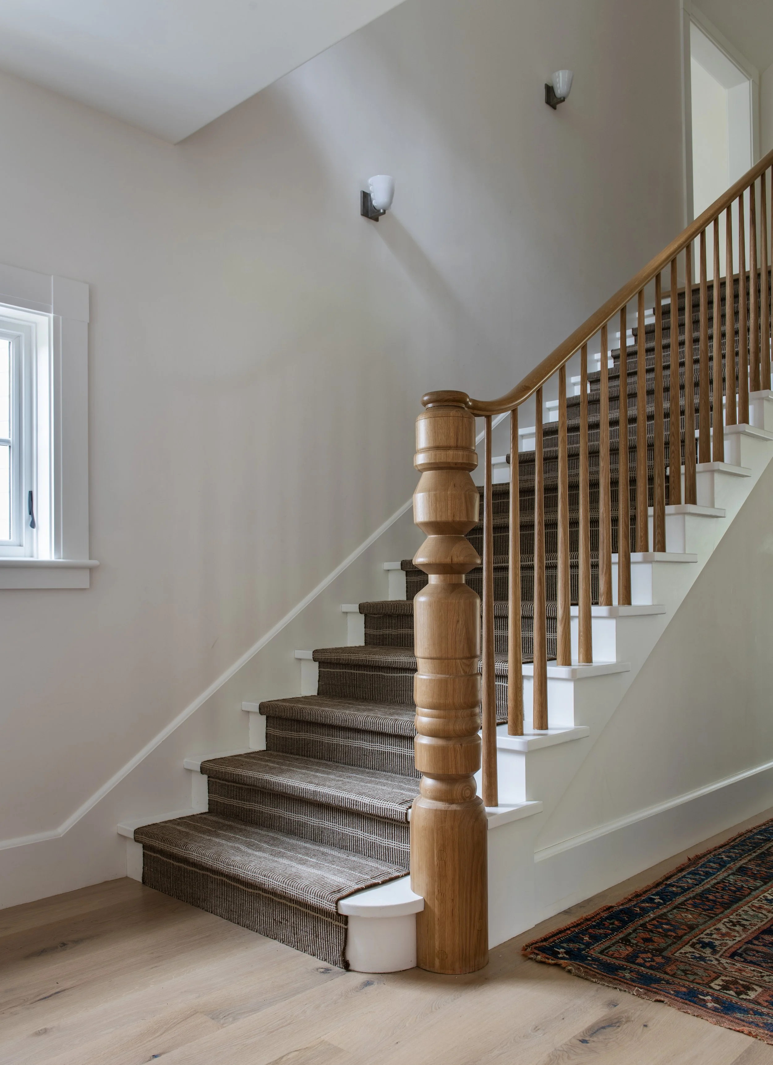 Interior view of a staircase with wooden handrail and striped carpeted stairs, a small window on the left, and a patterned rug on the floor near the stairs.