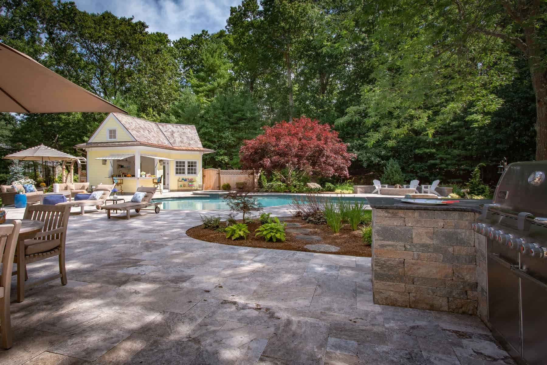 Backyard pool area with patio, lounge chairs, umbrellas, and a small yellow pool house surrounded by trees and lush greenery.