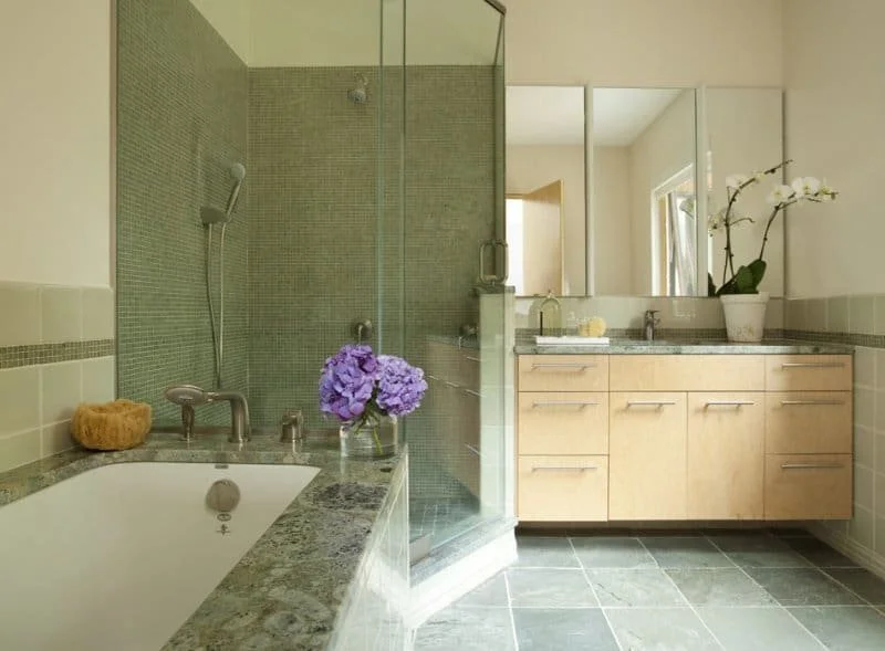 Modern bathroom with a glass-enclosed shower, a bathtub with a purple flower bouquet on the edge, a beige vanity with a large mirror, and a potted orchid plant.