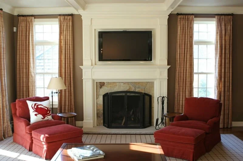 Living room with two red armchairs, a wooden coffee table, television above a fireplace, and large windows with beige curtains.
