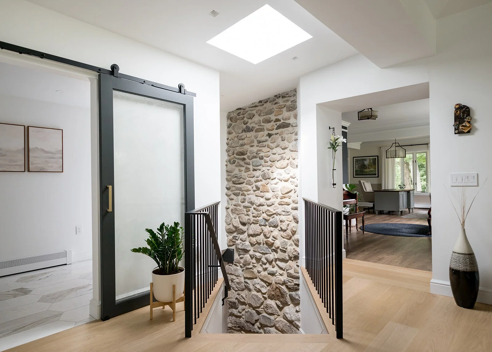 Interior view of a modern house entryway with a stone accent wall, black railing, potted plants, and open living room with large windows and greenery outside.