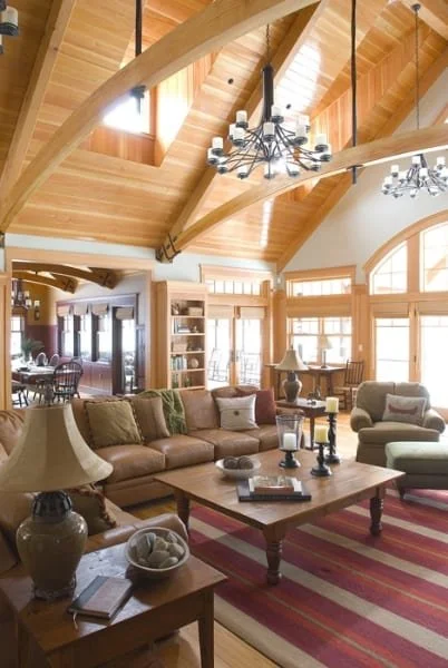 Living room with leather sofa, armchair, wooden coffee table with candles and decor, large windows, wooden ceiling with exposed beams, and hanging chandeliers.