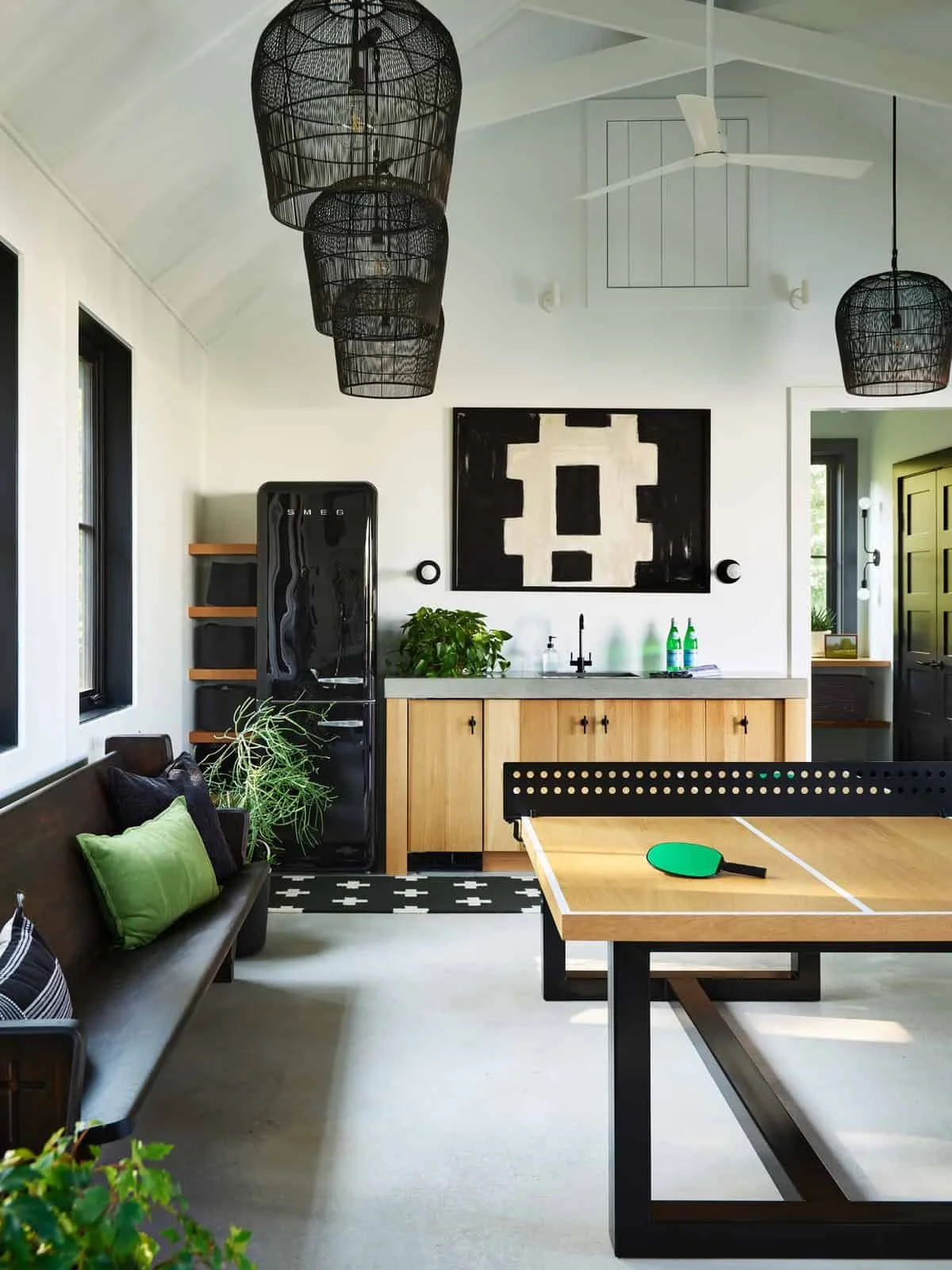Modern living room with black pendant lights, white ceiling fan, black refrigerator, wooden cabinet, plants, black and white artwork, and a ping pong table with a green paddle.
