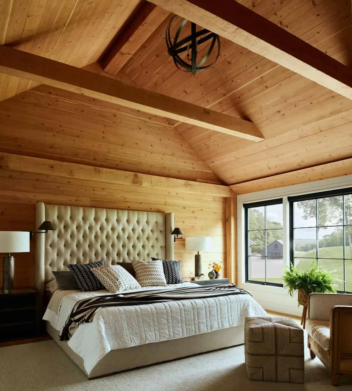 A cozy bedroom with wooden walls and a vaulted ceiling, featuring a large, padded headboard, a white bedspread, decorative pillows, a nightstand with a lamp and flower vase, and a window with a view of greenery outside.