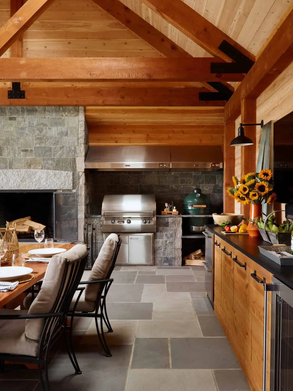 Interior of a rustic kitchen with wooden beams, stone walls, a stainless steel grill, a green ceramic grill, a wood dining table with beige cushioned chairs, and a countertop with a vase of sunflowers, fresh fruits, and kitchen utensils.