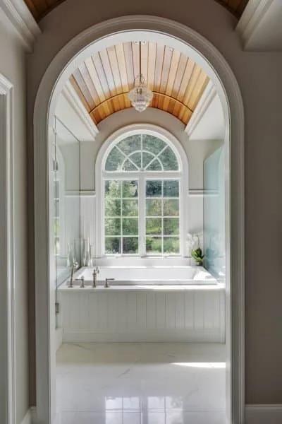 Bright bathroom with large arched window, bathtub, and wooden ceiling, decorated with a small plant and elegant fixtures.