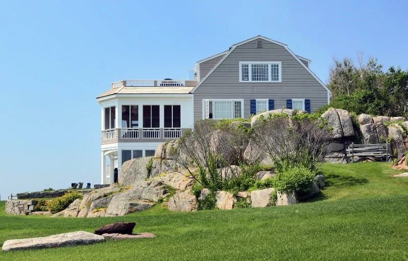 A large grey house with a front porch and multiple windows, situated atop a rocky hill with green grass and shrubs, under a clear blue sky.