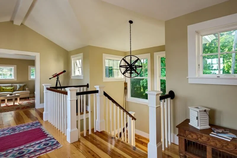 Bright upstairs landing of a house with cream walls, wooden floors, and multiple white-framed windows showing green foliage outside, decorated with a black modern chandelier, a telescope, and a wooden table with a small white lantern and papers.