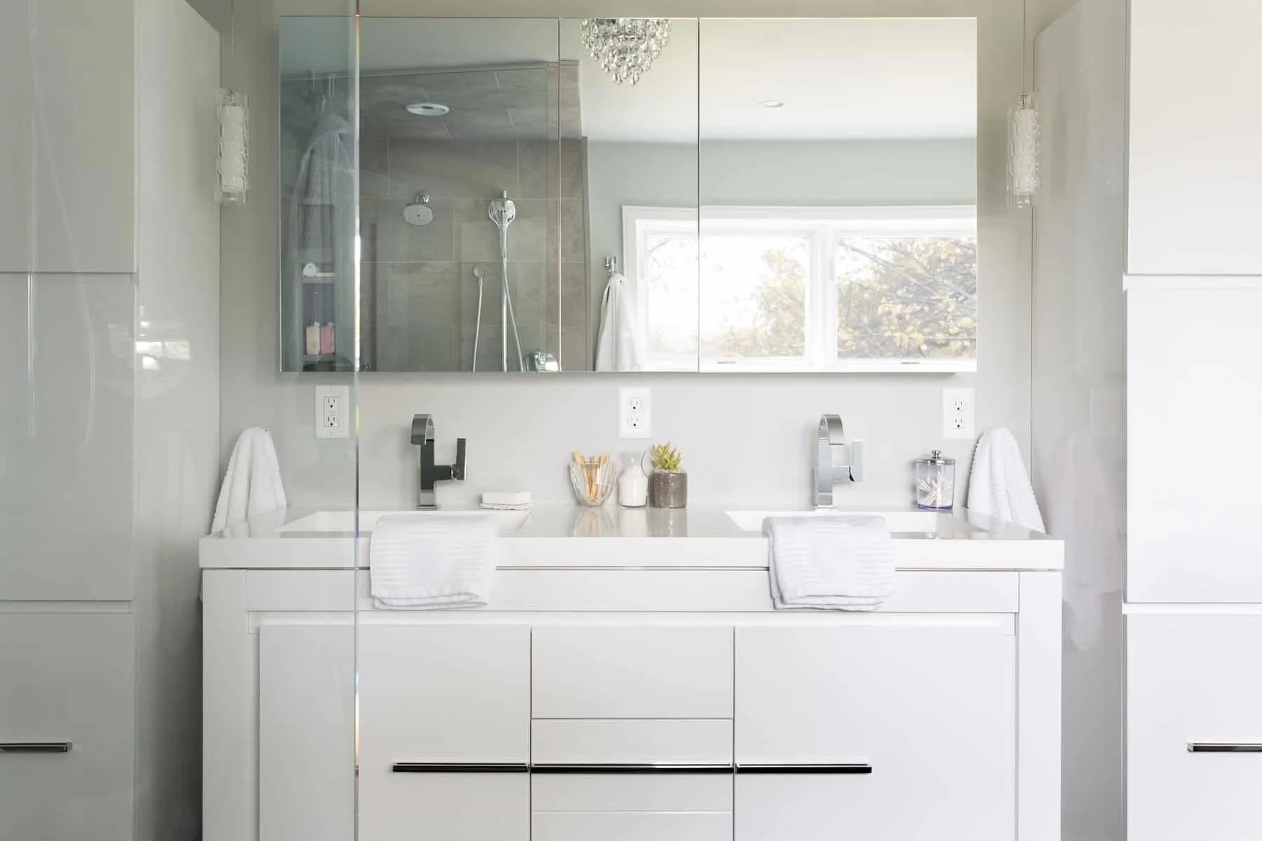 A modern white double bathroom vanity with two sinks, black faucets, and a large mirror above. The vanity has drawer storage with horizontal black handles, and towels hang on each side. Decor items like jars, a small plant, and brushes are on the cou