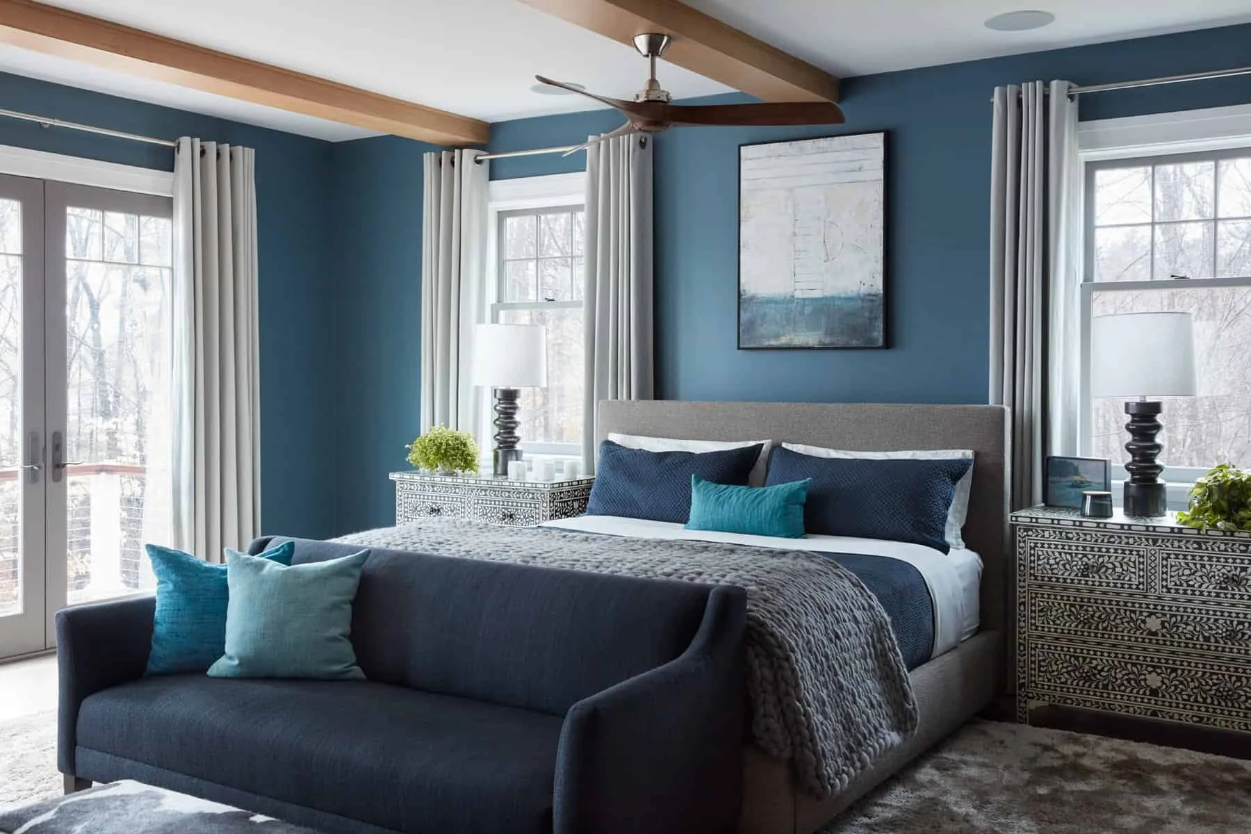 A bedroom with blue walls and large windows with white curtains, a bed with navy and turquoise pillows, and a cushioned bench in front with matching pillows. There are nightstands with lamps, plants, and decorative items, and a ceiling fan with woode