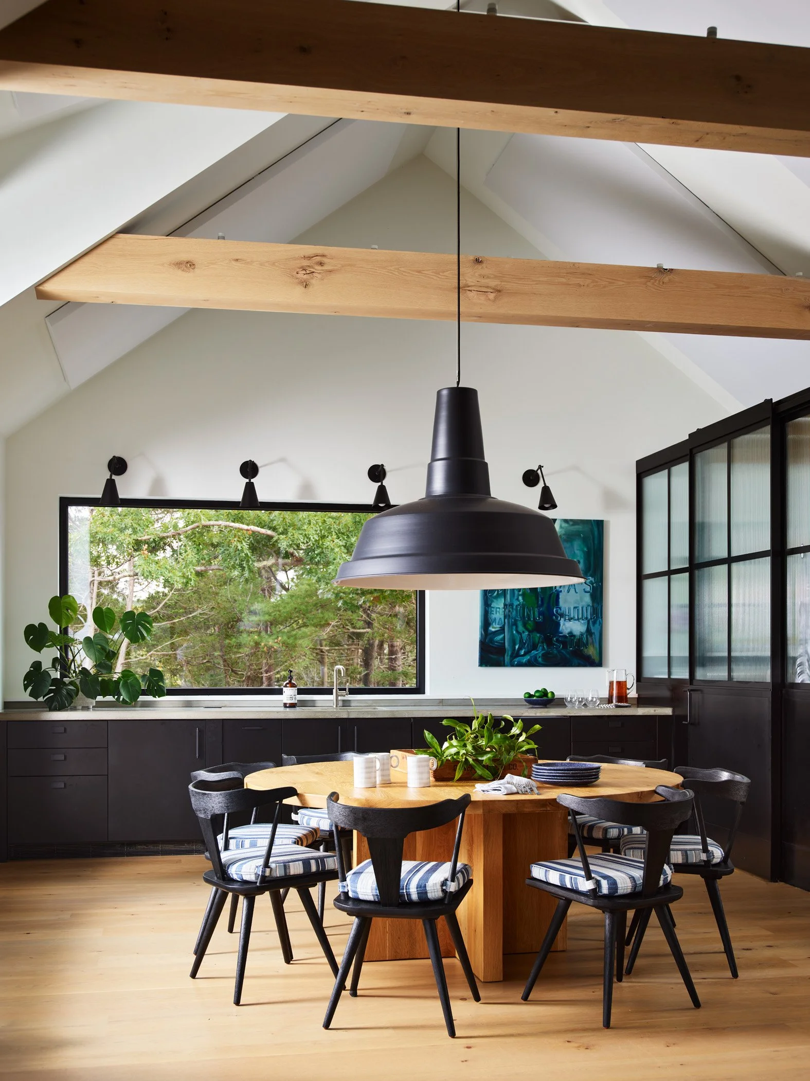 A modern kitchen with a round wooden dining table surrounded by eight black chairs with striped cushions. There is a large window showing green trees outside, a black pendant light hanging above the table, dark cabinets, and a black glass cabinet on 