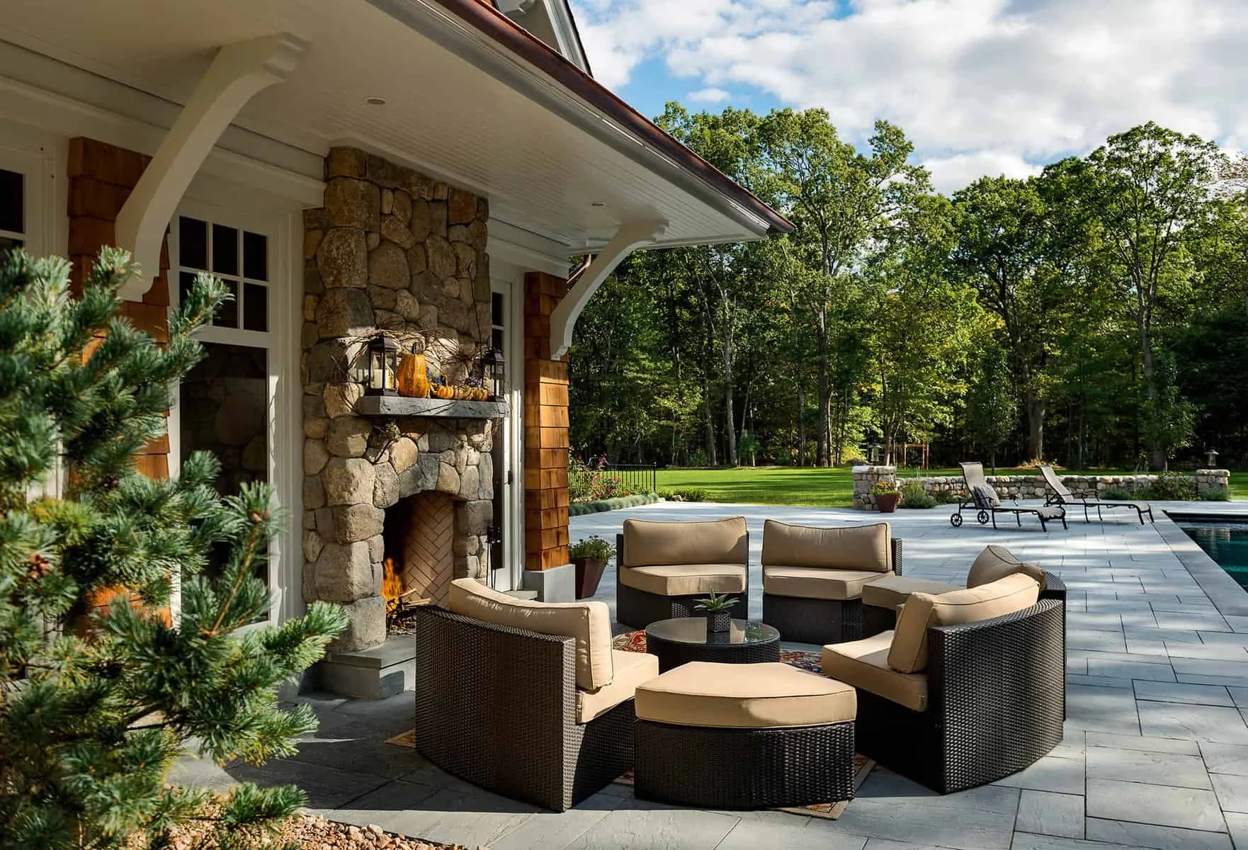 On a patio, wicker chairs with beige cushions surround a small black table, next to a stone fireplace with a mantel decorated with lanterns, pumpkins, and gourds. A fragment of a green pine tree is visible in the foreground. The background features a