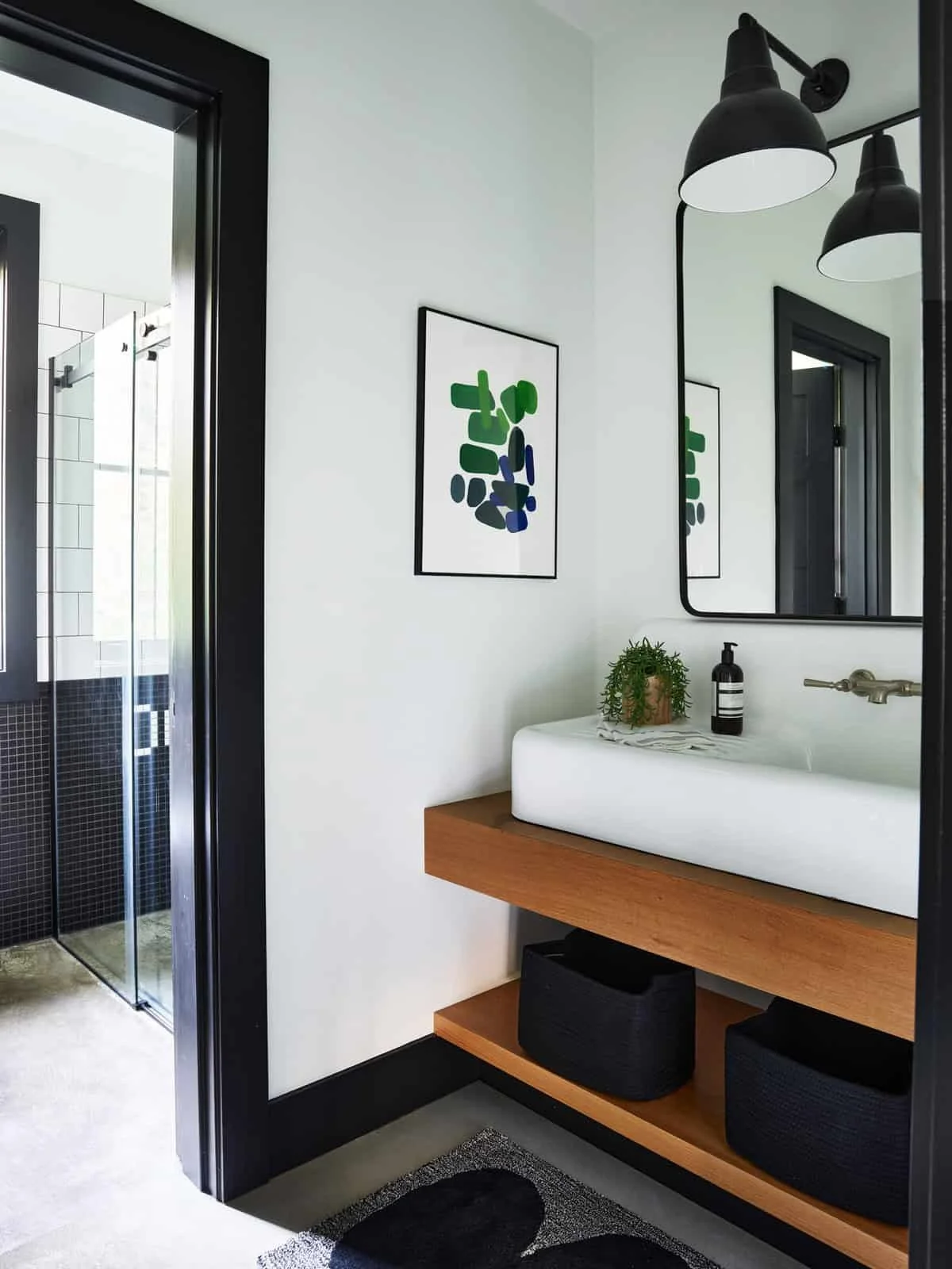 Modern bathroom featuring a wooden vanity with a vessel sink, black and white decor, a large mirror, black light fixtures, a framed artwork, a small green plant, and a black storage basket. A glass shower with white tiled walls and black accents is v