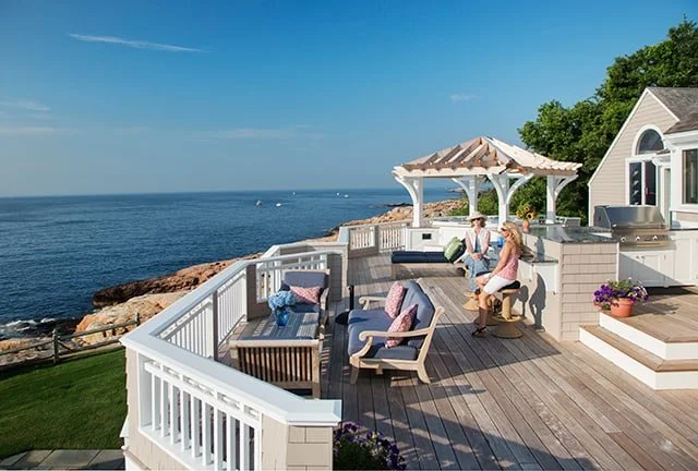 A spacious oceanfront deck with outdoor seating, a small gazebo, and a house overlooking the water with a rocky shoreline and blue sky.