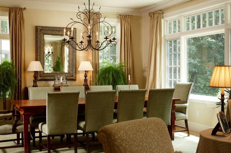 A dining room with a large wooden table, eight upholstered chairs, a chandelier, and large windows with beige curtains, surrounded by greenery outside.