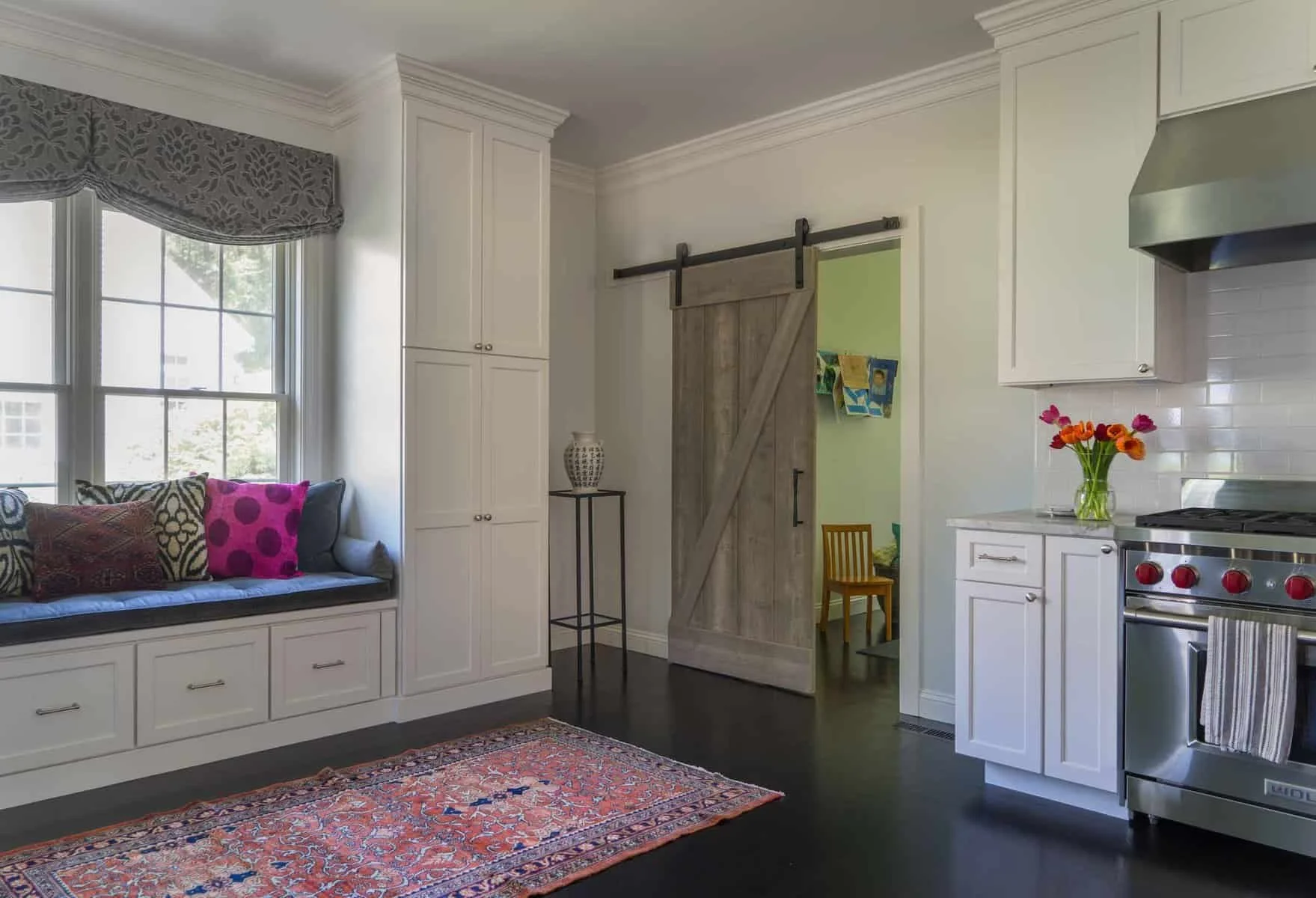 Interior of a kitchen with built-in window seat, white cabinets, sliding barn door, and stainless steel stove with a vase of colorful flowers on the counter.