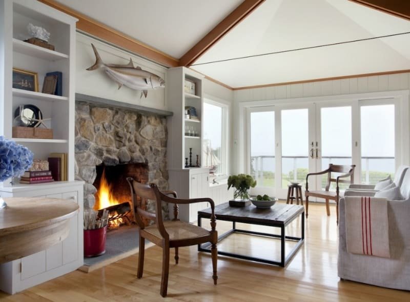 Living room with a stone fireplace, a mounted fish above it, and large glass doors leading outside. Furnished with wooden chairs, a white sofa, a black coffee table with a plant, and built-in shelves with decorative items.