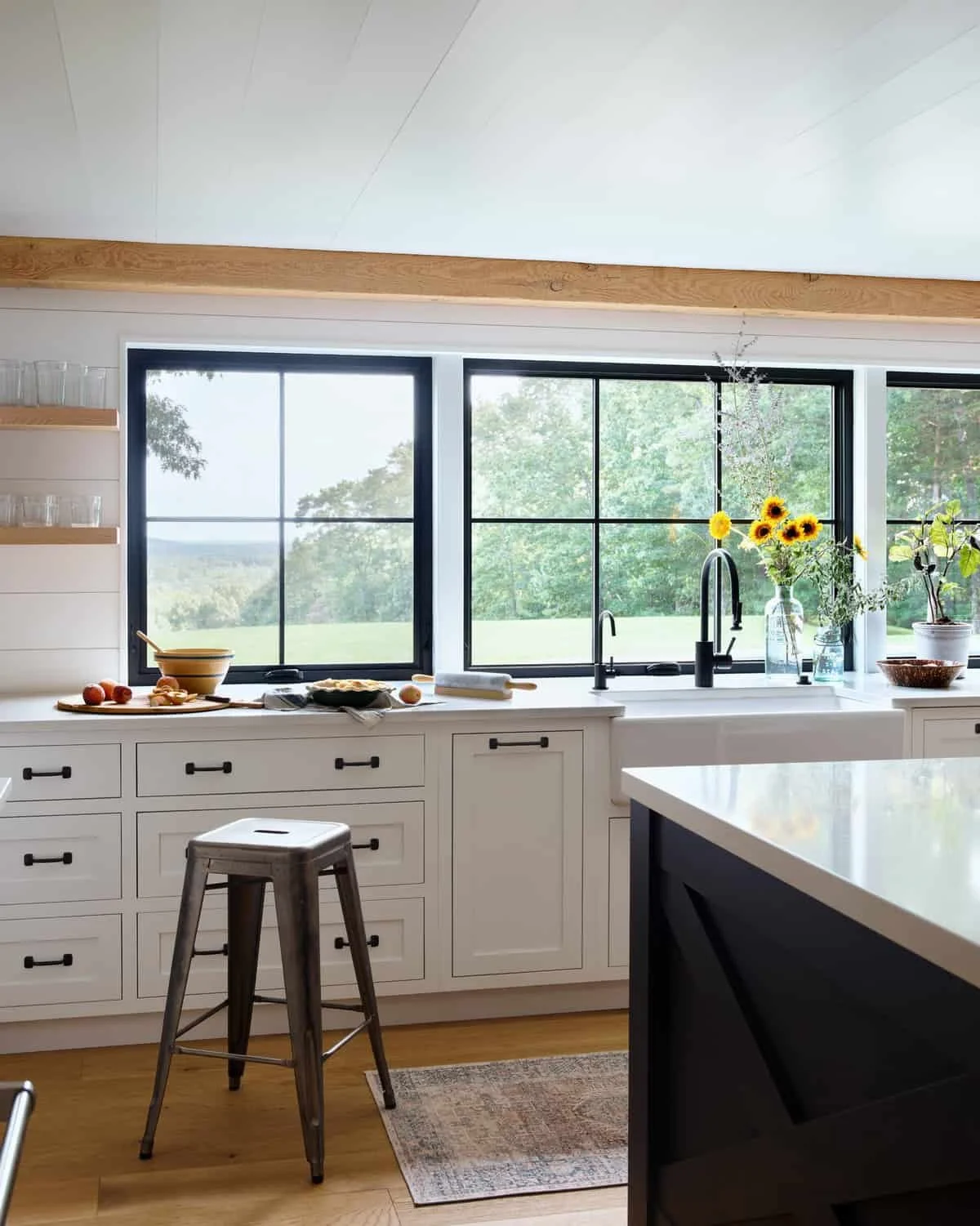 Modern kitchen with black-framed large windows, white cabinetry, a kitchen island, and a view of green trees outside. Decorated with vases of sunflowers and other plants, with a table and a bar stool in the foreground.