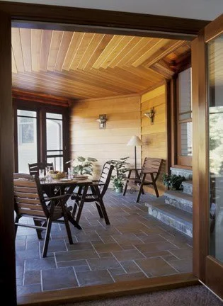 Indoor patio with wooden walls and ceiling, a dining table with chairs, potted plants, and sunlight coming through large windows.