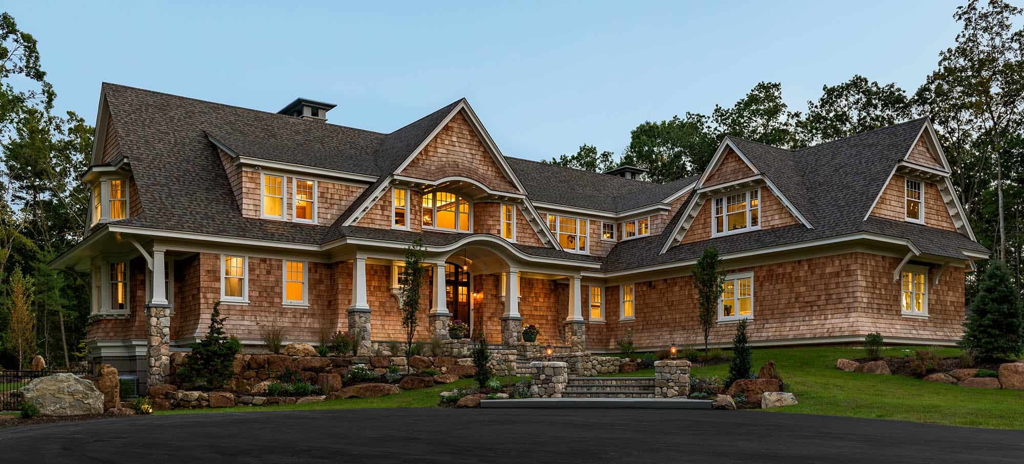 Large house with multiple gabled roofs, stone foundation, and wooden siding, illuminated from inside, surrounded by a landscaped yard with trees and rock decorations.