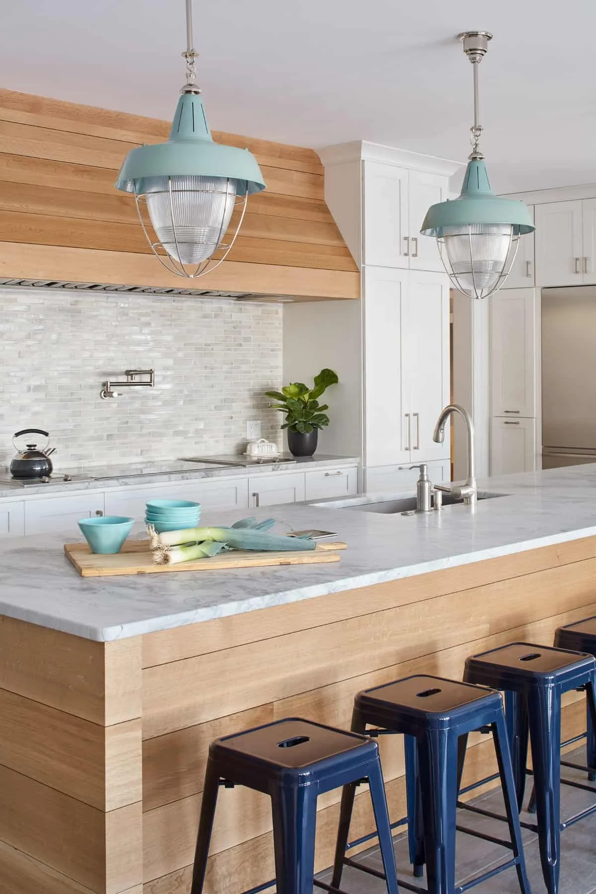 Modern kitchen with white cabinets, wooden accents, marble countertop island, two blue pendant lights, a black kettle, a plant, and blue bar stools.