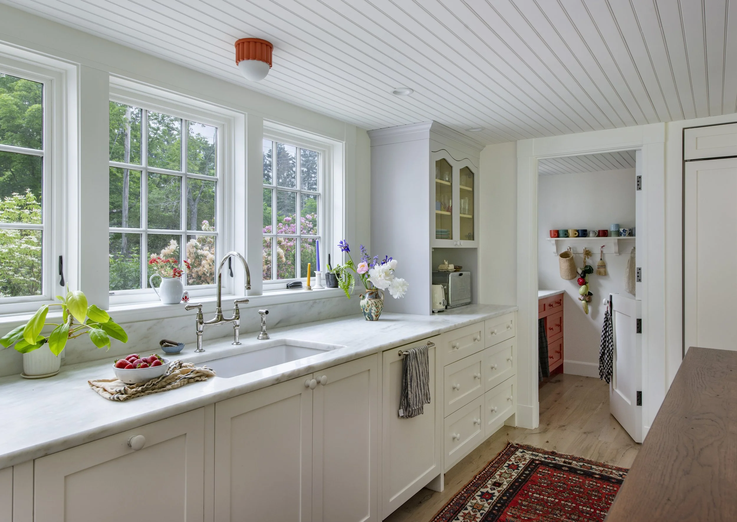 A bright kitchen with large windows, white cabinetry, a marble countertop, and a view of green trees outside. The countertop has a potted plant, a bowl of strawberries, and flowers in a decorative vase. A small red rug is on the wooden floor, and the