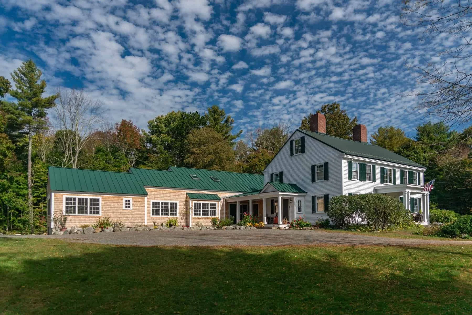 A white farmhouse with green shutters and a green metal roof, surrounded by trees with a partly cloudy sky overhead.