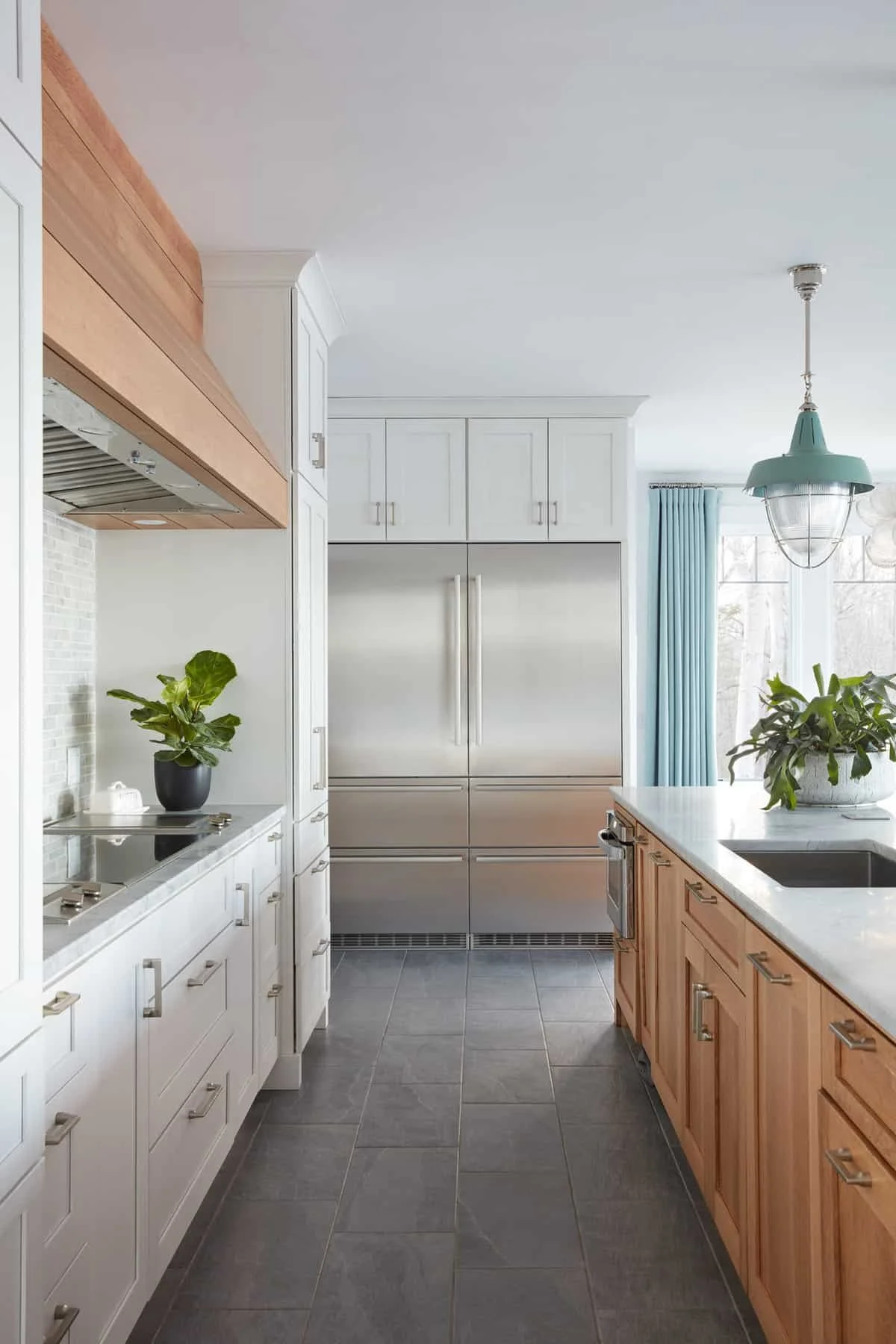 Modern kitchen with white cabinets, stainless steel refrigerator, wooden island with sink, gray tile floor, potted plants, blue curtains, and a hanging light fixture.